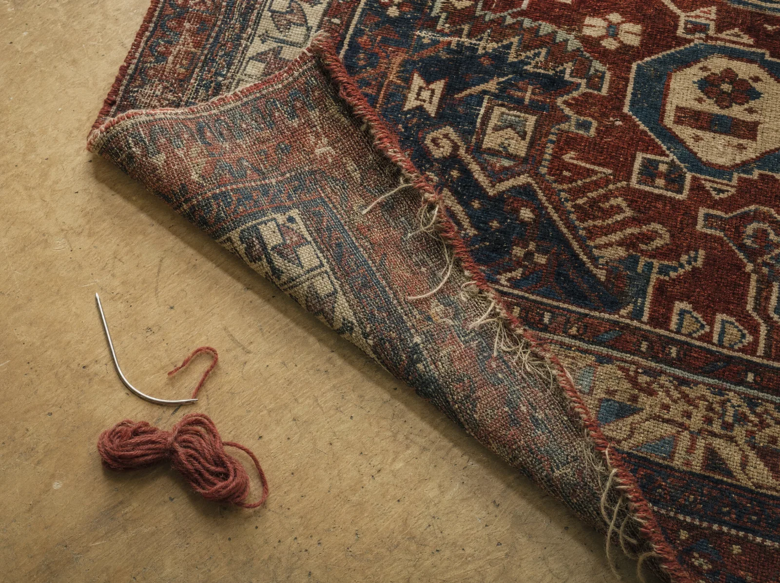 Lisière usée d'un tapis ancien, aiguille et laine rouge assortie posées à côté (illustration)