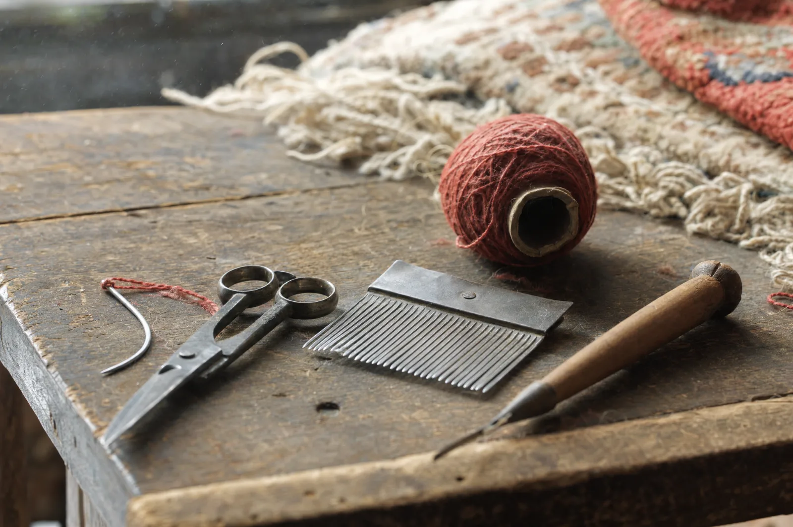 Outils de restauration de tapis sur un établi : aiguille courbe, peigne, ciseaux et bobine de laine rouge (illustration)